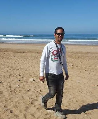 Mann mit Sonnenbrille und weißem Shirt spaziert am Sandstrand bei sonnigem Wetter mit Wellen im Hintergrund